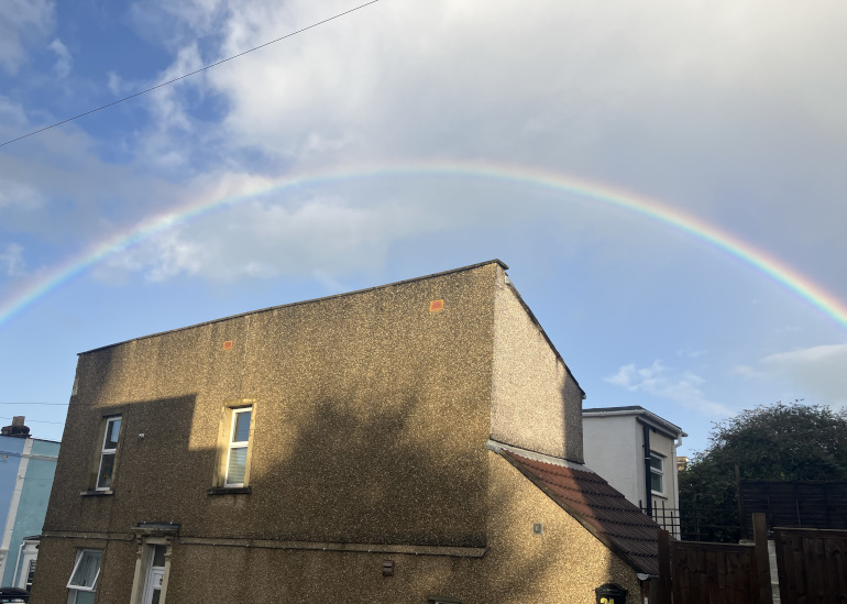 House arched over by a rainbow in a blue sky by Judy Darley